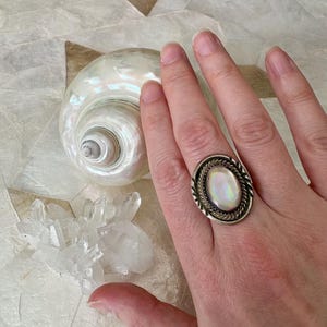 Pode incluir: Um anel de prata com uma pedra cabochão oval iridescente engastada numa borda de metal torcido. O anel é usado num dedo, posicionado perto de uma concha grande, lisa e nacarada, e de um grupo de cristais de quartzo transparentes e irregulares.