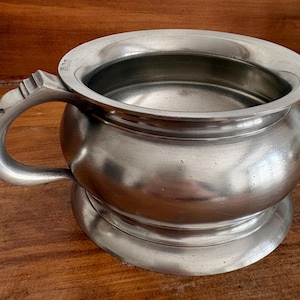 Op de afbeelding: Een zilverkleurige metalen kom met een brede, ronde opening en een enkele gebogen handgreep aan de zijkant. De kom heeft een glad, afgerond lichaam dat taps toeloopt naar een platte, ronde voet, met kleine stempelmarkeringen op de rand.