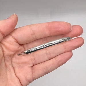 May include: A silver-colored tie clip with etched floral and geometric designs. The clip is held in a hand against a light background. The clip is approximately 2.5 inches long.