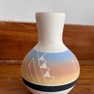 Peut inclure: Un vase en céramique blanc mat avec un corps arrondi et un col étroit. La partie centrale présente un motif dégradé passant du bleu à l'orange et au jaune, agrémenté de motifs géométriques de voiles gravés et d'une bande noire horizontale.