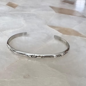 Peut inclure: Un bracelet manchette argenté avec des motifs tribaux gravés. Le bracelet ouvert est en métal brillant et repose sur une surface claire. Le design du bracelet présente des motifs géométriques répétés.