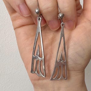 Puede incluir: Un par de pendientes de plata con un diseño geométrico. Los pendientes están hechos de un metal fino y delicado y tienen una forma larga y delgada.