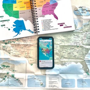 Może przedstawiać: Zbiór map i smartfon wyświetlający mapę Stanów Zjednoczonych. Otwarty notatnik w oprawie spiralnej z mapą stanów USA. Widoczna jest również złożona mapa systemu parków narodowych.