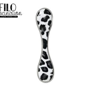 Puede incluir: Un reposacucharas decorativo con un estampado de manchas blancas y negras, que recuerda al pelaje de un dálmata. El reposacucharas tiene un borde plateado y una forma alargada única. El texto "FILO fascination" está en la esquina superior izquierda.