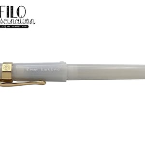 May include: A pearlescent white Pilot Kakuno fountain pen with gold-colored accents. The pen has a gold clip and a gold band around the cap. The text "FILO fascination" is above the pen.