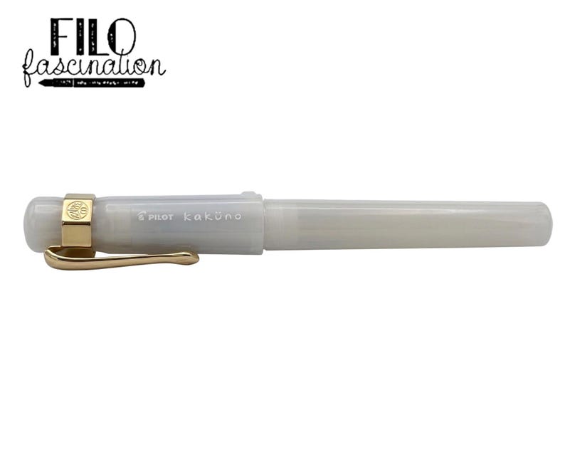 May include: A pearlescent white Pilot Kakuno fountain pen with gold-colored accents. The pen has a gold clip and a gold band around the cap. The text "FILO fascination" is above the pen.