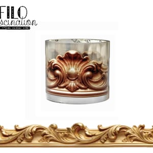 Puede incluir: Un portavelas cilíndrico de cristal con un patrón de relieve en oro metálico y volutas ornamentadas. El texto FILO fascination está impreso en negro en la esquina superior izquierda. Debajo se sitúa un borde decorativo dorado a juego.