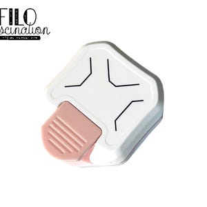 Puede incluir: Una herramienta redondeadora de esquinas blanca y rosa con líneas negras. La herramienta tiene una palanca rosa y el texto "FILO fascination" en escritura negra.