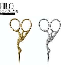 Puede incluir: Tijeras de bordar doradas y plateadas con un diseño decorativo de grulla. Las tijeras tienen orificios grandes para los dedos y una punta afilada. El texto "FILO fascination" está en la esquina superior izquierda.