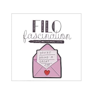 Könnte beinhalten: Ein pinkfarbener Emaille-Pin mit einem offenen Umschlag, einem Herz und den Worten "YOU'RE DOING A GREAT JOB". Darüber ein schwarzer Stift und die Worte "FILO fascination".