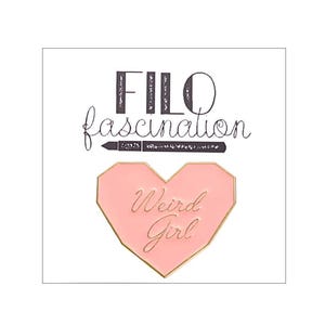 Puede incluir: Un broche de esmalte en forma de corazón de color rosa claro con bordes dorados. El texto 'Weird Girl' está grabado en cursiva dorada en el centro. Sobre el broche, el texto 'FILO fascination' está impreso en negro con una línea horizontal.