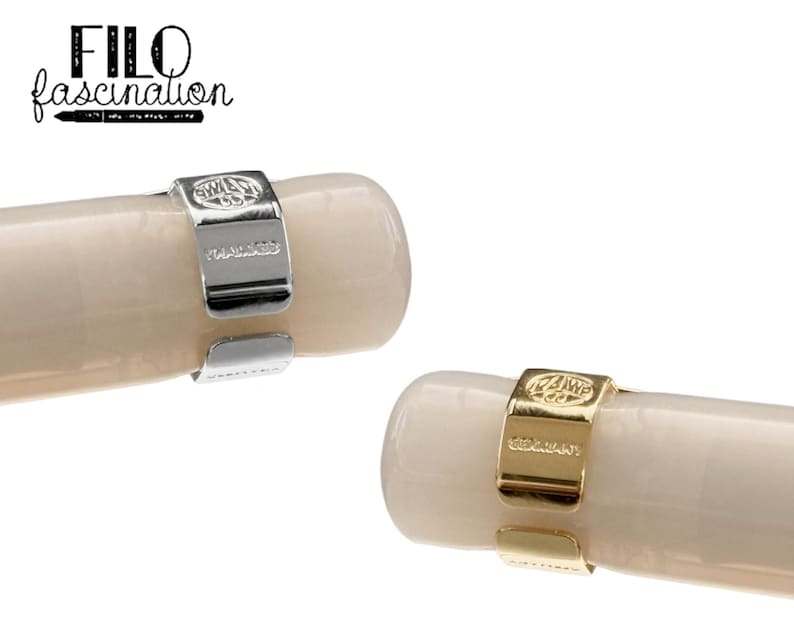 May include: Close-up of two pen clips, one silver and one gold, each encircling a cream-colored pen barrel. The silver clip is stamped with "KAWECO GERMANY". The gold clip also has "KAWECO GERMANY" stamped on it. The text "FILO fascination" is in the upper left corner.