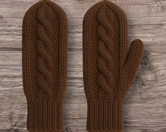 Puede incluir: Un par de mitones de punto marrón con un diseño de trenzado. Los mitones son de un color marrón cálido y terroso y parecen estar hechos de un hilo suave y texturizado. El patrón de trenzado recorre el centro de cada mitón.