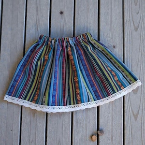 May include: A colorful, striped skirt with a white lace trim. The skirt features vertical stripes in blue, green, red, and orange. The skirt is made of a lightweight fabric and has an elastic waistband.