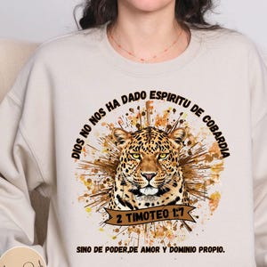May include: Beige sweatshirt featuring a leopard graphic with text in Spanish. The leopard is surrounded by a brown and gold burst design. The text includes "DIOS NO NOS HA DADO ESPIRITU DE COBARDIA" and "2 TIMOTEO 1:7".