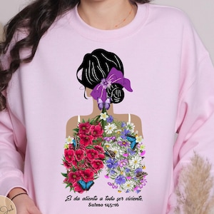 May include: A light pink sweatshirt featuring a graphic design of a woman's silhouette with floral lungs and butterflies. The design includes red, purple, and white flowers. Text at the bottom reads "El da aliento a todo ser viviente, Salmo 145:16."
