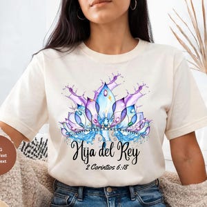Peut inclure: T-shirt crème avec un motif de fleur de lotus aquarelle dans des tons violets et bleus. Le texte "Hija del Rey" et "2 Corintios 6:18" est écrit en script noir. C'est un vêtement décontracté.