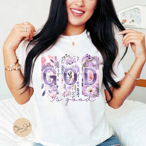 May include: White t-shirt featuring the phrase "GOD is good" in a floral design. The letters are purple and filled with pink and purple flowers. The shirt has a crew neck and short sleeves.