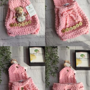 May include: A pink knitted backpack with a flap and drawstring closure. The backpack has wooden letter beads spelling "NERGIS" and a small teddy bear ornament. The bag is made of a chunky knit material.