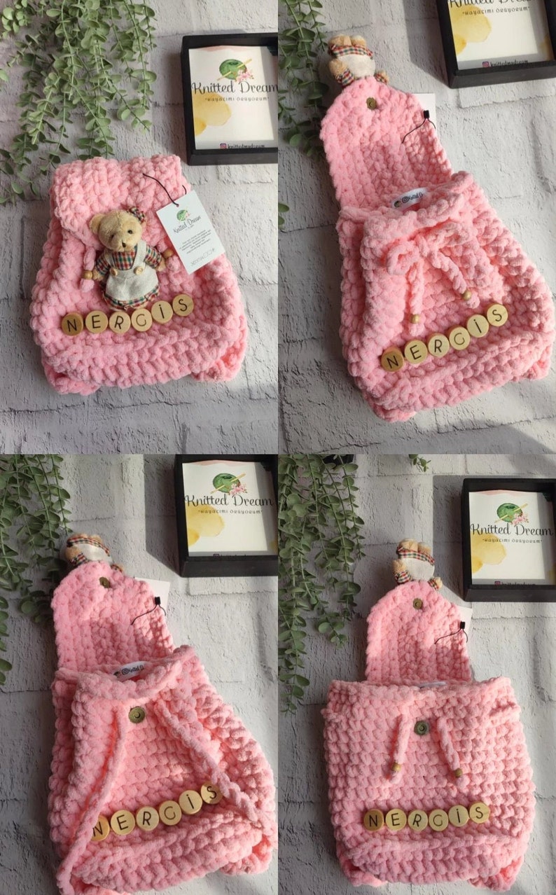 May include: A pink knitted backpack with a flap and drawstring closure. The backpack has wooden letter beads spelling "NERGIS" and a small teddy bear ornament. The bag is made of a chunky knit material.
