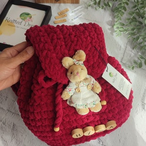 May include: A burgundy knitted backpack with a flap closure and wooden bead details. A small stuffed bunny wearing a floral dress and bow is attached. The backpack has a tag that says "Knitted Dream".