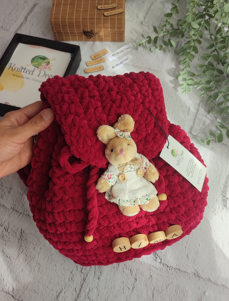 May include: A burgundy knitted backpack with a flap closure and wooden bead details. A small stuffed bunny wearing a floral dress and bow is attached. The backpack has a tag that says "Knitted Dream".