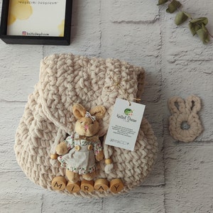 May include: A cream-coloured, chunky-knit backpack with a plush bunny attached. The bunny wears a floral dress and a bow. Wooden letters spell "MIRAY" at the bottom. A matching bunny-shaped teether is also present.