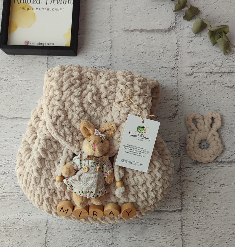 May include: A cream-coloured, chunky-knit backpack with a plush bunny attached. The bunny wears a floral dress and a bow. Wooden letters spell "MIRAY" at the bottom. A matching bunny-shaped teether is also present.