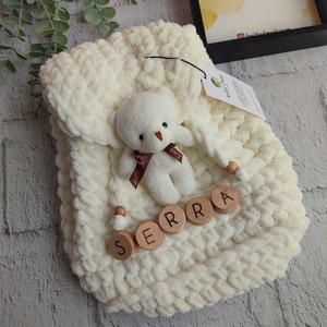 May include: A cream-coloured, chunky knit baby blanket with a small white teddy bear and wooden letter blocks spelling "SERRA". The bear has a brown ribbon. The blanket is folded and has a tag attached.
