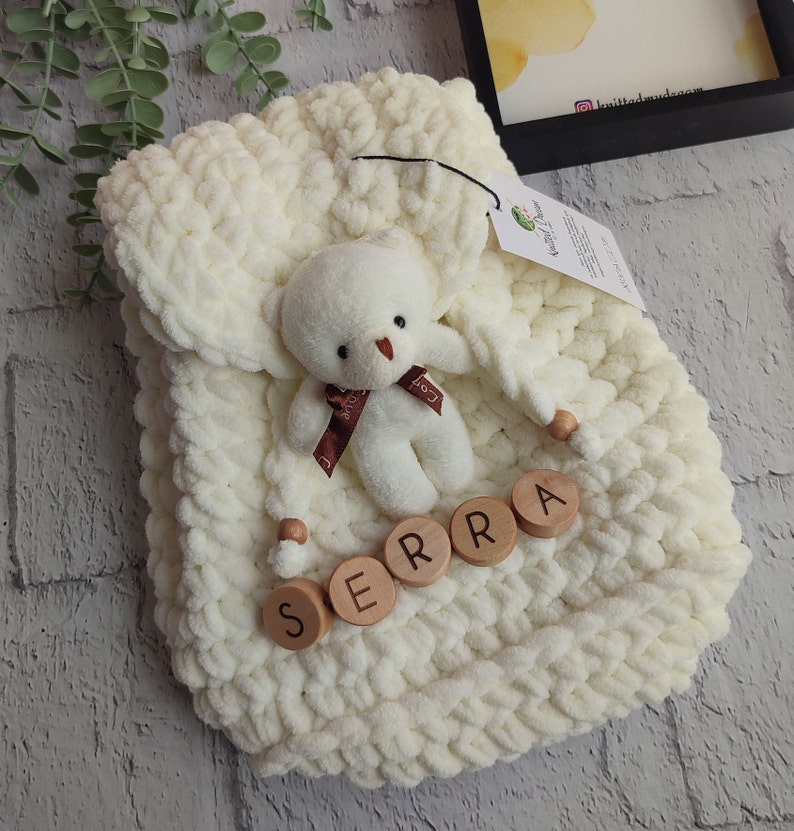 May include: A cream-coloured, chunky knit baby blanket with a small white teddy bear and wooden letter blocks spelling "SERRA". The bear has a brown ribbon. The blanket is folded and has a tag attached.