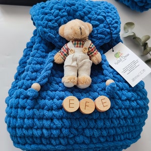 May include: A handmade blue knitted backpack with wooden detailing and a small teddy bear wearing dungarees. The backpack features a drawstring closure and wooden buttons with the letters "EFE". A tag with text is attached.
