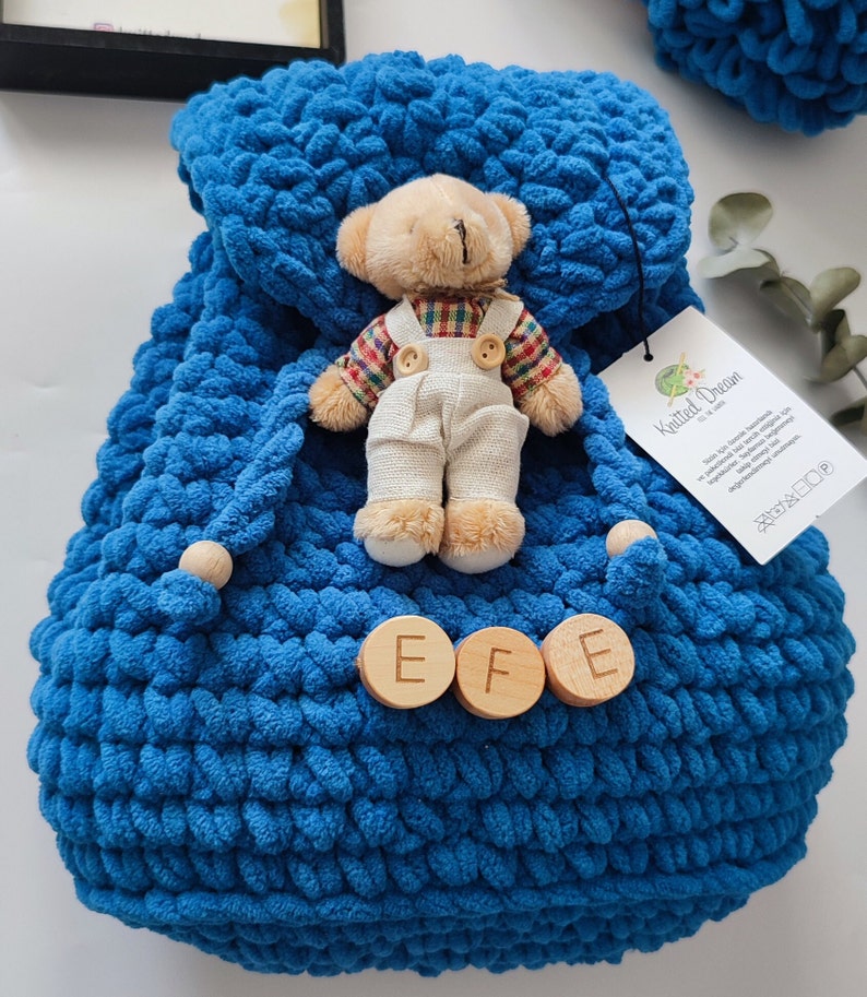 May include: A handmade blue knitted backpack with wooden detailing and a small teddy bear wearing dungarees. The backpack features a drawstring closure and wooden buttons with the letters "EFE". A tag with text is attached.