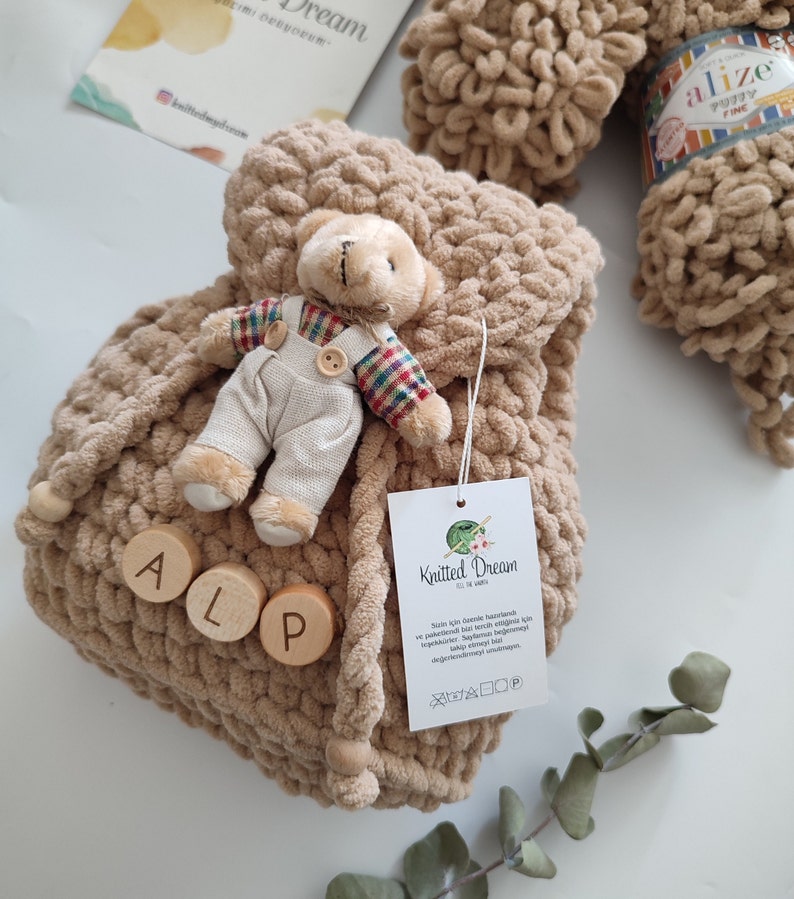 May include: A beige, knitted drawstring bag with wooden beads and the letters "ALP". A small teddy bear in overalls is attached to the bag. A ball of Alize Puffy Fine yarn and a tag with the "Knitted Dream" logo are also visible.