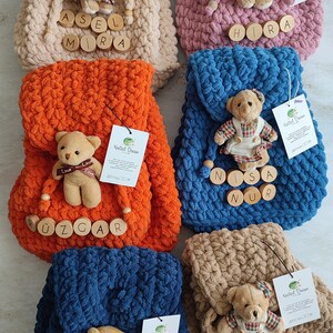 May include: Several handmade, plush backpacks in shades of beige, pink, orange, blue, and brown. Each backpack is adorned with a small teddy bear and wooden name tags. These backpacks are designed for children.