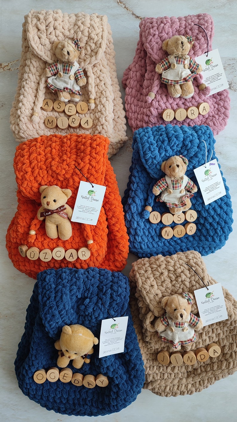 May include: Several handmade, plush backpacks in shades of beige, pink, orange, blue, and brown. Each backpack is adorned with a small teddy bear and wooden name tags. These backpacks are designed for children.