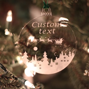 Könnte beinhalten: Ein transparenter Weihnachtsbaumschmuck aus Glas mit einer weißen Silhouette einer Winterszene, darunter ein Haus, Bäume und der Weihnachtsmann in einem von Rentieren gezogenen Schlitten. Der Schmuck trägt außerdem den Text "Custom text", der darauf graviert ist.