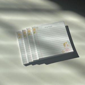 May include: A stack of lined notepads with floral accents in yellow, pink, and green. The word "notes" is printed at the top of the notepad. The notepads are stacked on a white surface, with shadows cast across them.