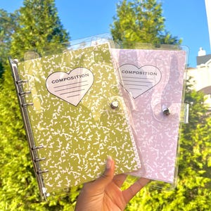 May include: Two composition notebooks with clear plastic covers. One notebook is light green with white splatters, the other is light purple with white splatters. Each notebook has a heart-shaped label with the word "COMPOSITION".