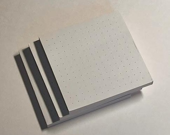 May include: A stack of square, dot grid paper notebooks. The pages are white with small, evenly spaced dots. The edges of the pages are visible, showing the thickness of the stack. The notebooks are on a light-colored surface.