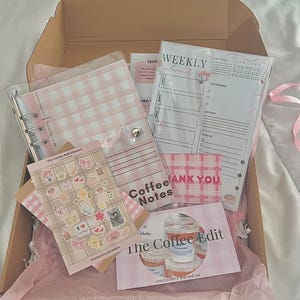 May include: An open cardboard box filled with pink tissue paper and stationery items. Includes a pink and white gingham binder, weekly planner sheets, stickers, and cards with text "Coffee Notes" and "The Coffee Edit".