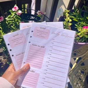 May include: A stack of pink and white planner pages with a daily overview, to-do list, and note sections. The pages have a quote by Audrey Hepburn. The pages are held in front of a background of flowers and greenery.