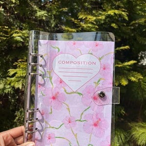 May include: A clear plastic binder with a pink floral design and the word "COMPOSITION" in a heart shape. The binder has a silver ring mechanism and a clear plastic cover.