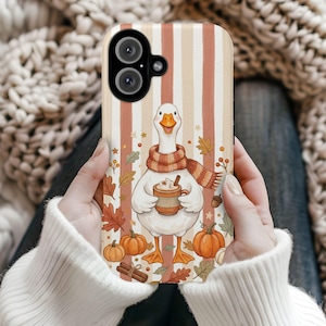 May include: A phone case featuring a white goose wearing a striped scarf and holding a mug of hot cocoa. The background has vertical beige and orange stripes, decorated with autumn leaves, pumpkins, cinnamon sticks, and small stars.