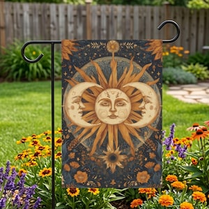 May include: A garden flag featuring a celestial design with a sun and two crescent moons. The artwork is in shades of gold and cream against a dark blue background, with floral accents. The flag is displayed on a black metal stand in a garden setting.