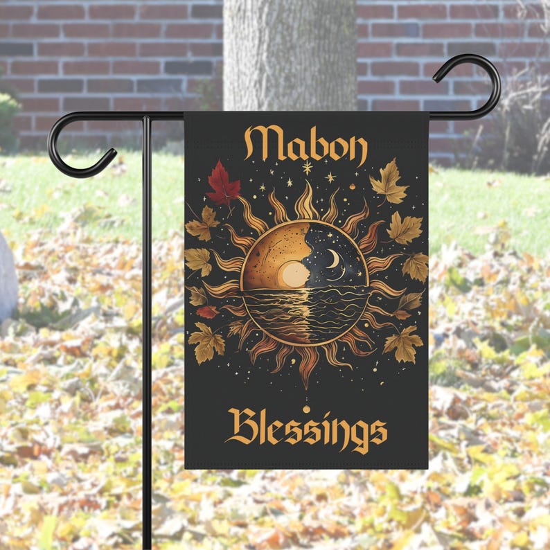 Garden flag with a stylized sun and moon over rippling water, encircled by autumn leaves and stars. The text reads “Mabon Blessings.”