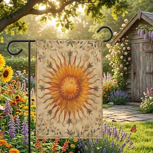 May include: A decorative garden flag featuring a stylized sun design in shades of orange and yellow. The flag is displayed on a black metal stand, set against a backdrop of a lush garden with flowers and a wooden shed.