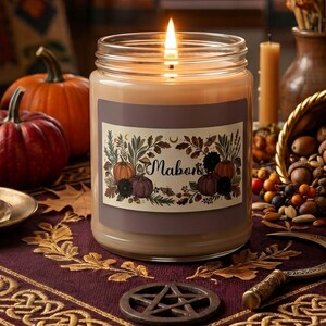 May include: A clear glass jar candle with a lit wick and light tan wax. The label features the text Mabon surrounded by illustrations of pumpkins, autumn leaves, and crescent moons. The candle sits on a dark purple cloth with gold embroidery.