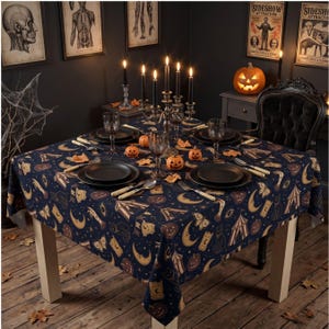 May include: A square tablecloth with a dark blue background featuring a repeating pattern of yellow crescent moons, orange pumpkins, black cats, moths, and circus tents. The table is set with black plates, silverware, and a silver candelabra.