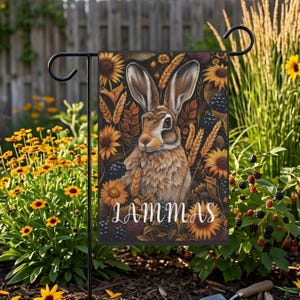 May include: A garden flag featuring a detailed illustration of a brown rabbit surrounded by sunflowers, wheat, and blackberries. The word "LAMMAS" is written in white script at the bottom. The flag is displayed on a black metal stand in a garden setting.