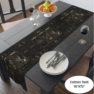 May include: A black cotton twill table runner measuring 16 inches by 72 inches, featuring a repeating tarot card design with gold-toned celestial motifs, moon phases, and intricate illustrations. The runner is laid flat on a dark wooden table.